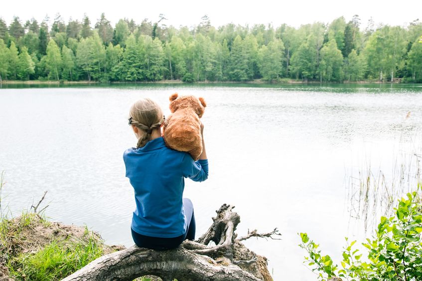 Girl holding stuffed teddy bear over shoulder while looking at the lake