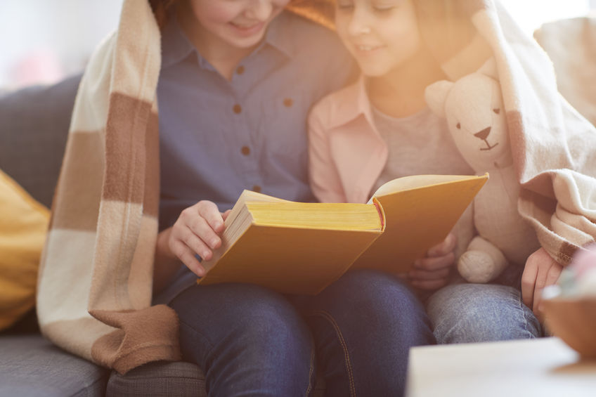Closeup portrait of two sisters reading book under blanket lit by warm sunlight, copy space