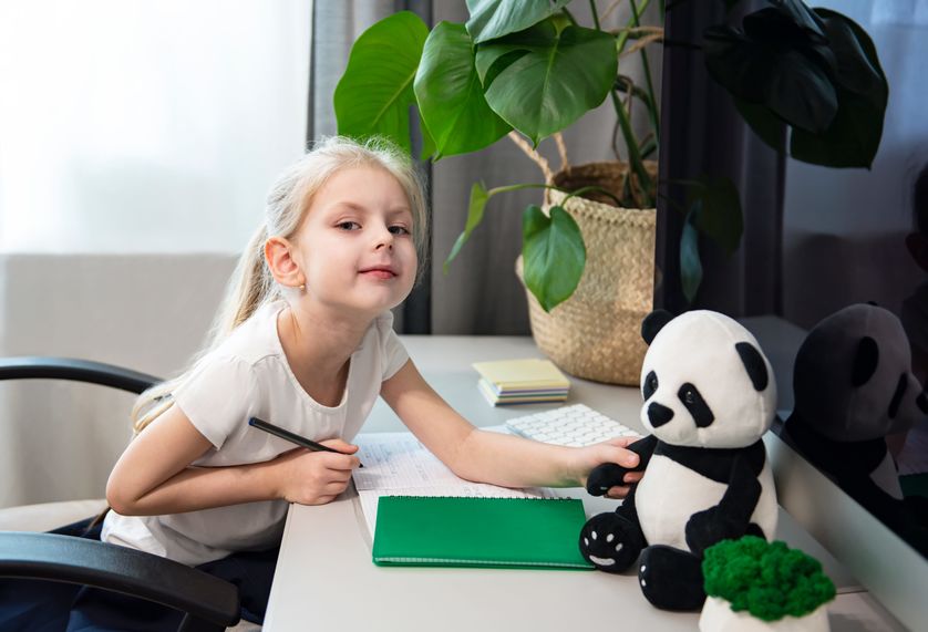 Little girl with blond hair sits at a table and does homework with stuffed animal panda.